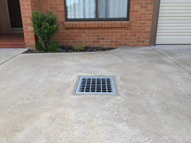 Concrete driveway with a square drain grate beside a brick house in Mount Druitt