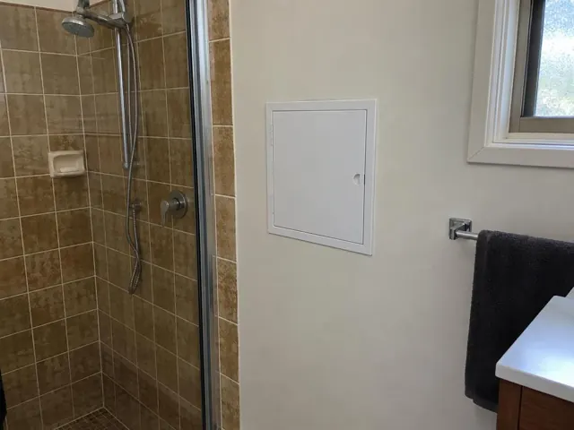Small access panel on a painted bathroom wall beside a tiled shower and towel rail in Woodcroft